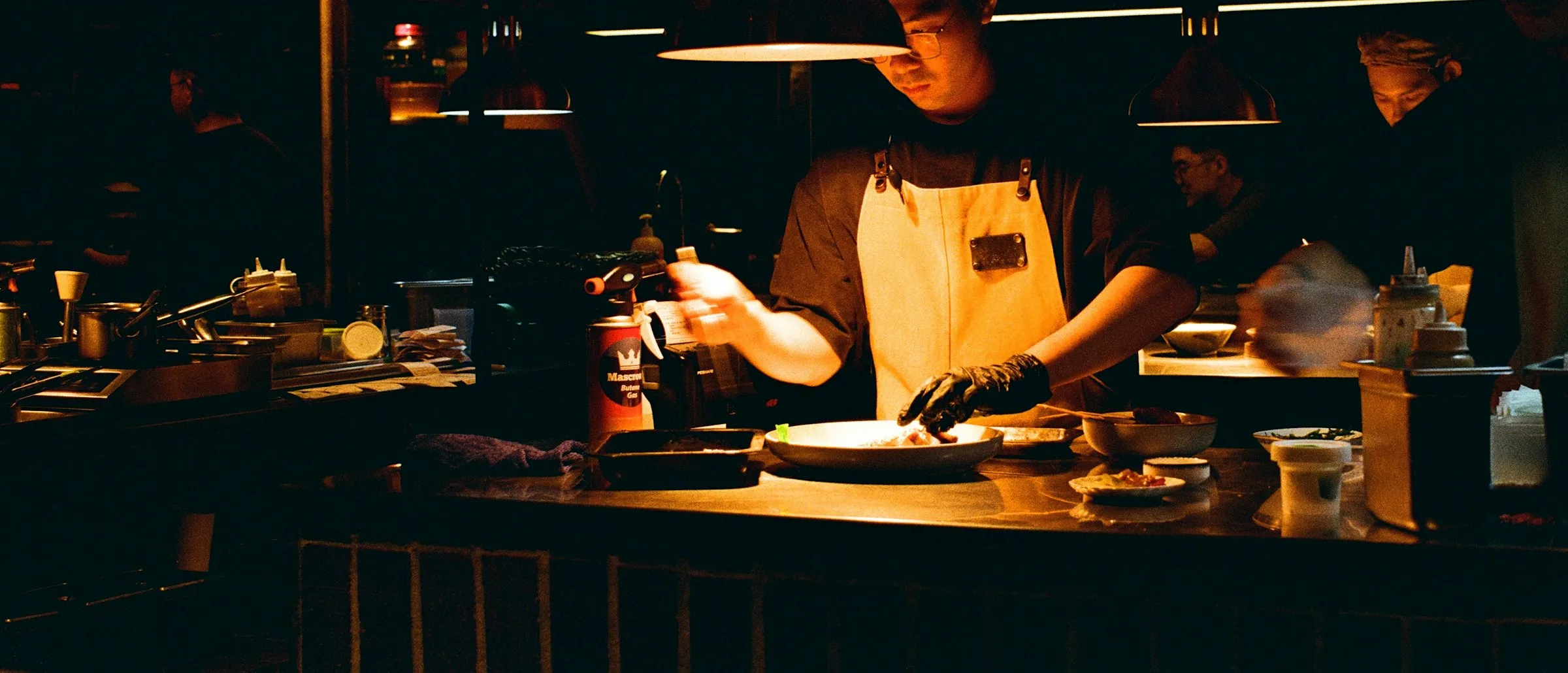 A chef plating at the pass of a dim, warmly lit open kitchen