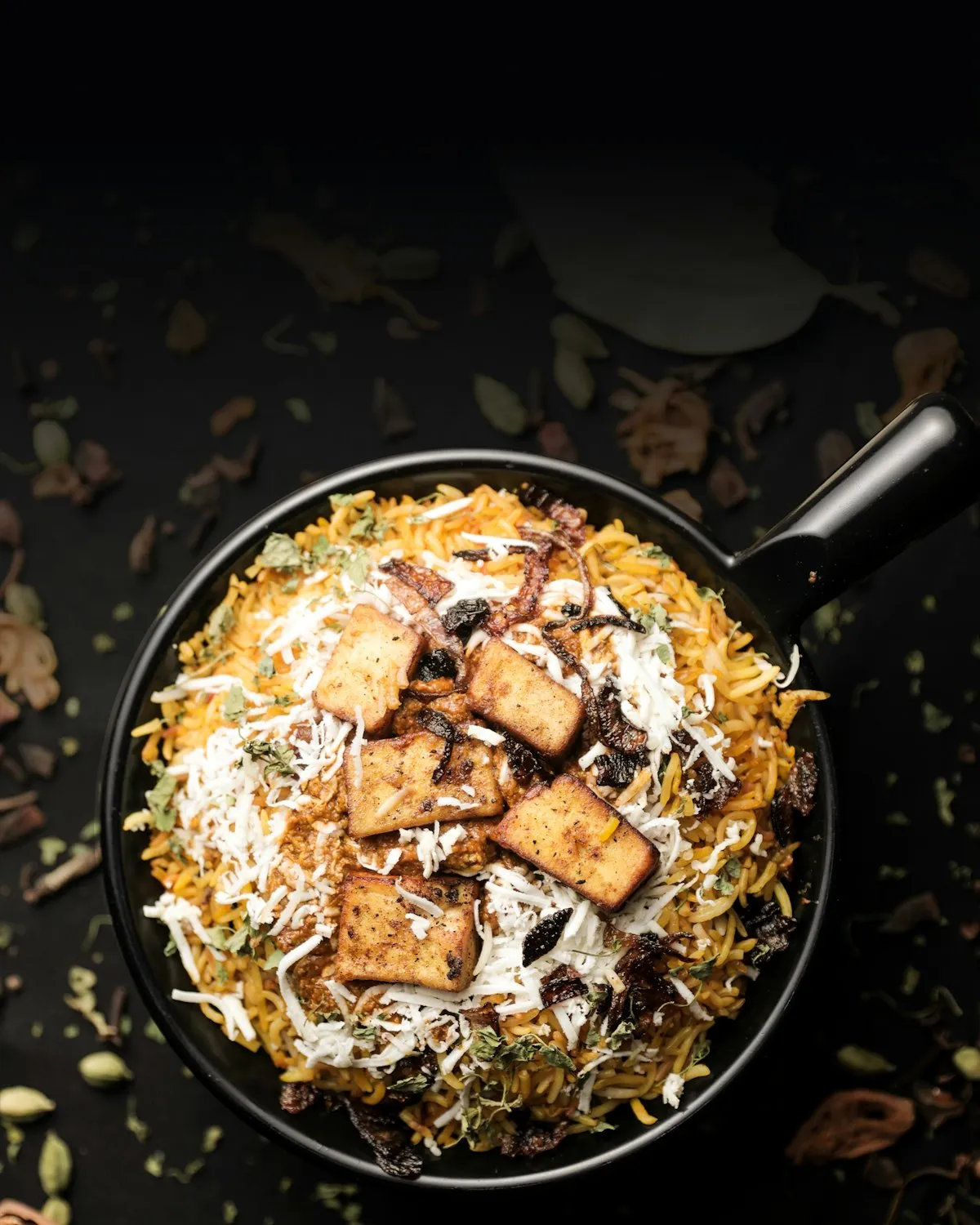Biryani in a black pan seen from above, topped with golden paneer and crisp onions