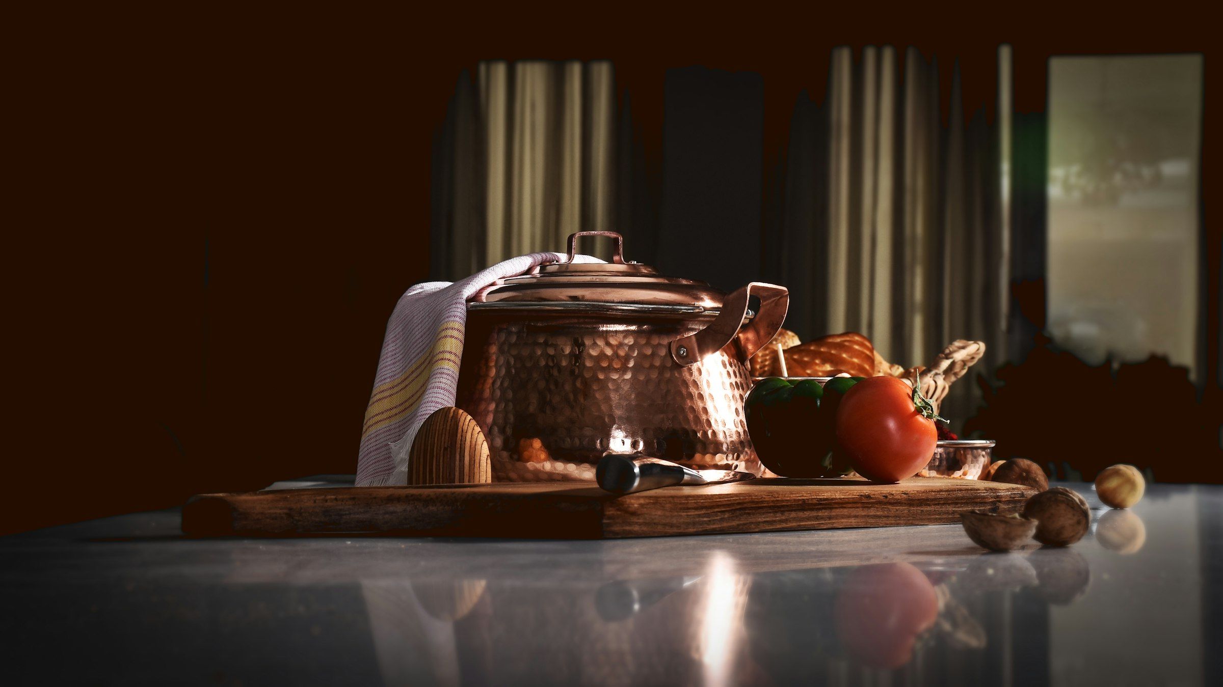 A hammered copper pot and fresh tomatoes on a wooden board in a dim, warm room