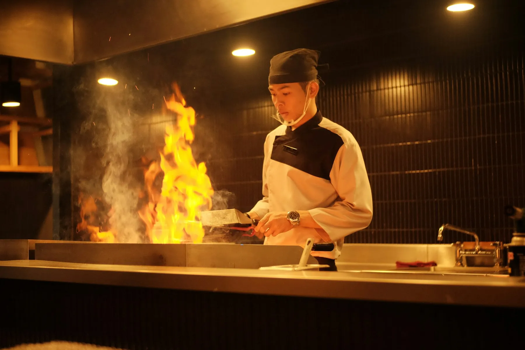 A chef working over a tall flame at the grill in a warmly lit kitchen