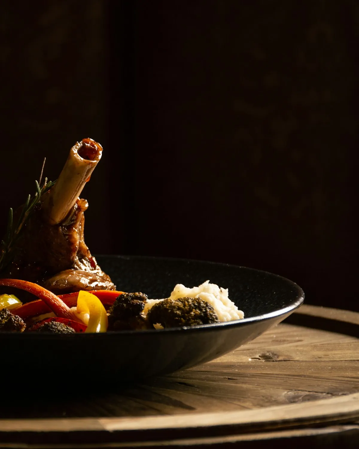 A slow-braised lamb shank in a dark bowl with roasted vegetables