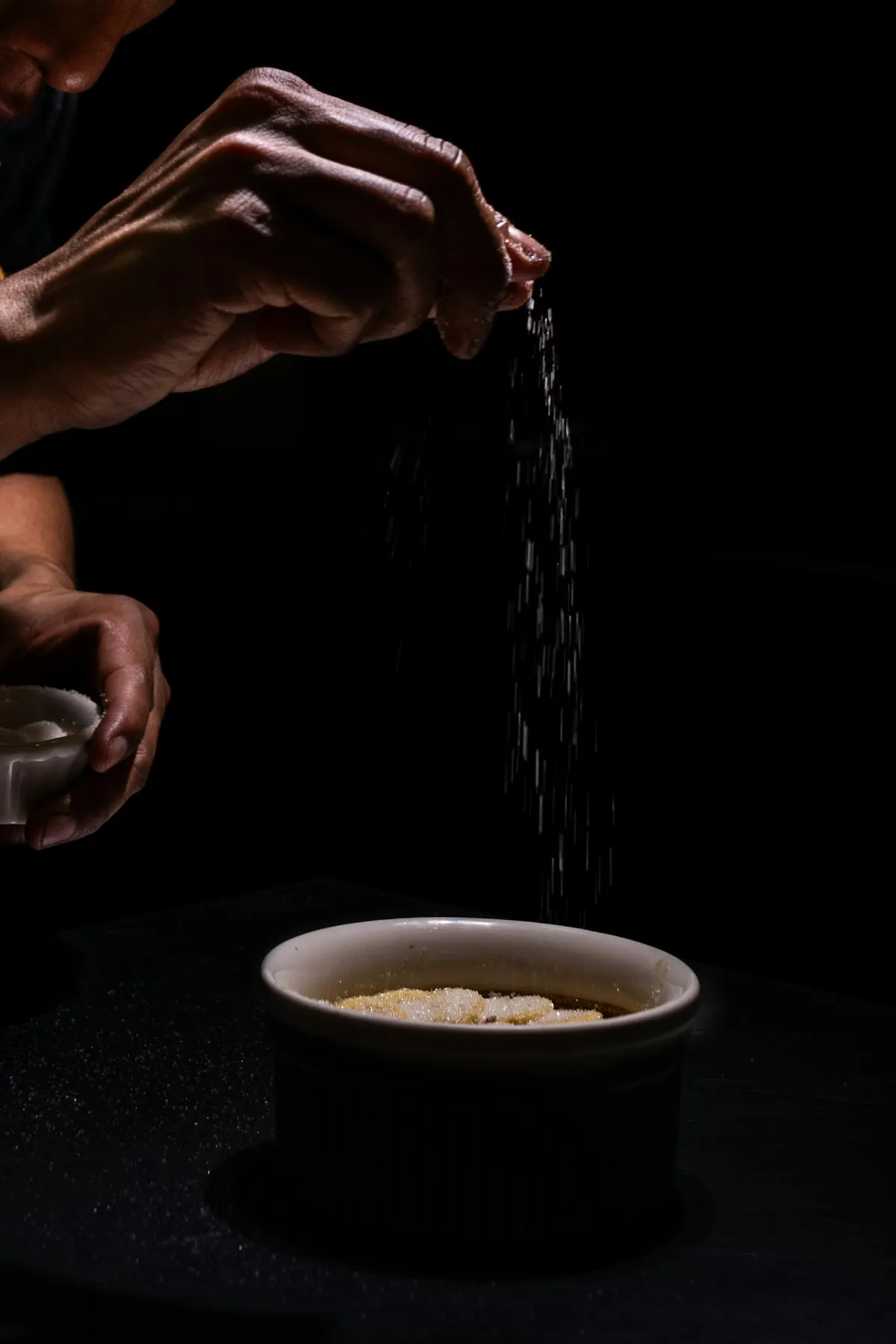 Hands in low light scattering a careful pinch of seasoning over a small dish