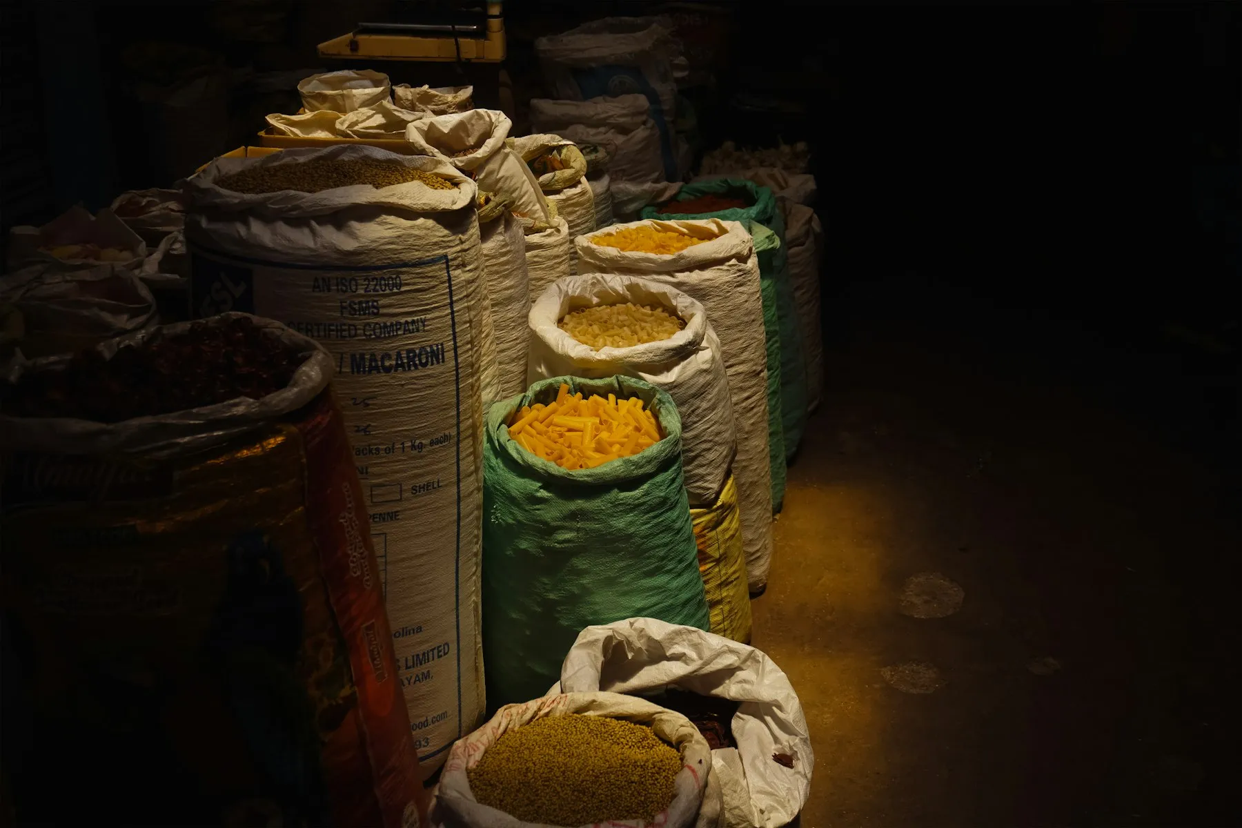 Open sacks of grain and ground spice in a dim storeroom, lit by a single warm lamp